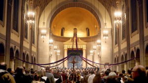 A cross bearer walks down the aisle of a crowded St. Joseph Memorial Chapel while the choir sings in the background.